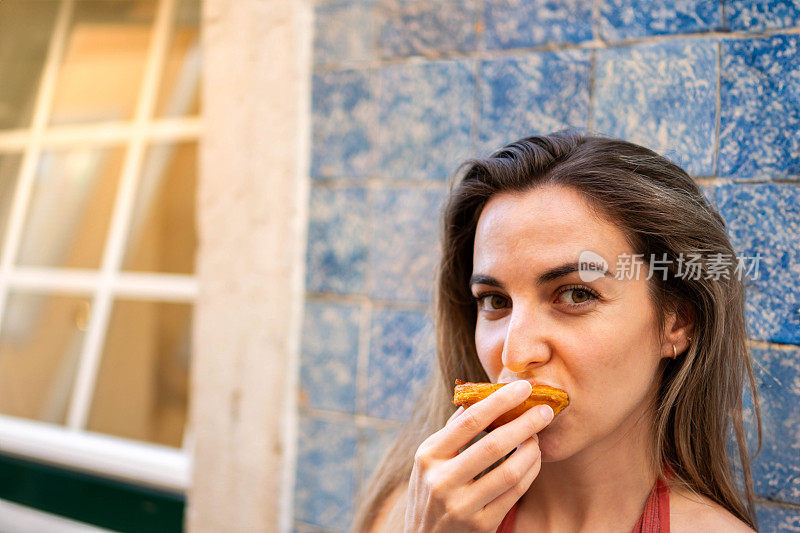 特写，侧视图的年轻女子品味传统的葡萄牙粉彩de Nata对一个充满活力的蓝色瓷砖墙，水平拍摄在日光下，体现了简单的快乐和文化遗产的本质